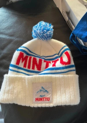 Vit Minttu mössa med blå tofs - Säljer en vit stickad Minttu mössa med blå och röda detaljer, samt en stor blåvit tofs på toppen. Mössan har bred uppvikt kant med Minttu-logga framtill och blå ränder runt om. Perfekt för kalla dagar och snygg att ha på vintern.
