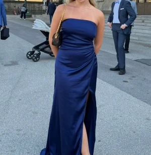 Balklänning - Säljer en marinblå axelbandslös långklänning i satin. Hög slits framtill och stilren design utan ärmar. Perfekt för t.ex balen💕 Inga defekter, ganska liten i storleken