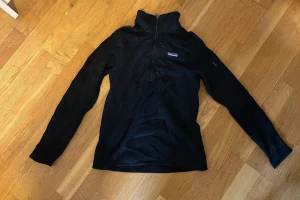Svart Patagonia half zip tröja - Svart långärmad tröja från Patagonia med half zip och hög krage. Perfekt för dig som gillar outdoor och vill ha en stilren look. Materialet är mjukt och lätt, troligen polyester, och tröjan har en normal passform. Diskret Patagonia-logga på bröstet.