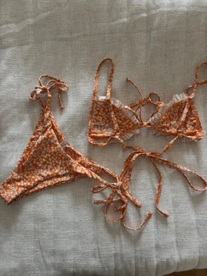 Orange blommig bikini med volang - Ny utan prislapp! Orange bikini från med vita små blommor och volangdetaljer på toppen. Både överdel och underdel har snörning och knyt i sidorna. Vadderade.