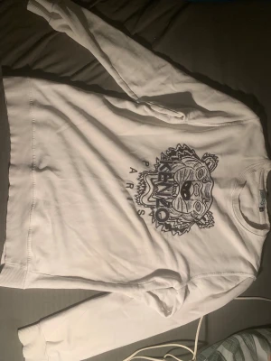Vit Kenzo sweatshirt med tigertryck XL men passar m-s - Säljer en vit sweatshirt från Kenzo i storlek XL med det ikoniska broderade tigerhuvudet och texten 'Kenzo Paris' på bröstet. Tröjan har ribbade muddar vid hals, ärmslut och nederkant. Perfekt för dig som gillar streetwear och snygga detaljer.