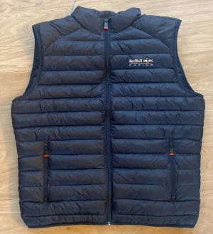 Svart dunväst Red Bull Racing x Pepe Jeans - Helt nya oanvänd,. Snygg svart dunväst från Red Bull Racing x Pepe Jeans med horisontella quiltade paneler, dragkedja framtill och två fickor med dragkedja. Västen har diskreta röda detaljer och logga på bröstet. 