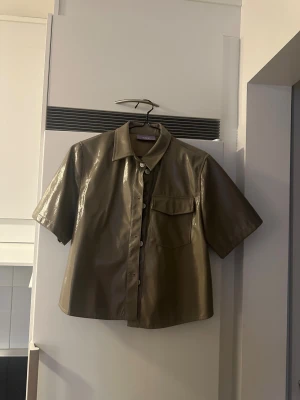Olivgrön kortärmad skjorta i skinn - Snygg olivgrön skjorta från Nelly i skinnimitation med korta ärmar och klassisk krage. Skjortan har en bröstficka och knappar framtill. Perfekt för dig som gillar en edgy och trendig look.