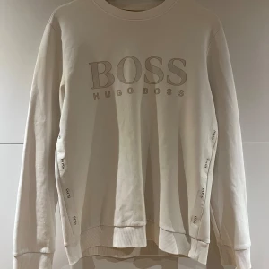 Vit sweatshirt från Hugo Boss - Snygg vit sweatshirt från Hugo Boss med broderad logga framtill och diskreta Boss-detaljer längs ärmarna. Klassisk rund halsringning och ribbade muddar. Perfekt för en clean och stilren look.