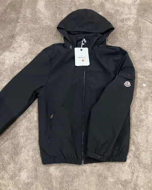 Svart vindjacka från Moncler med huva - Svart vindjacka från Moncler med huva, dragkedja framtill och fickor med zip. Jackan har Moncler-logga på ärmen och coolt serietryck på insidan. Elastisk nederkant och ärmslut med knapp. Perfekt för dig som gillar stilren och sportig look.
