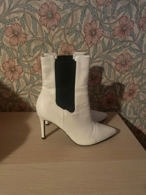Vita klackar  - Snygga vita stövletter från ESHOES med spetsig tå och hög klack. Skorna har svarta elastiska paneler på sidorna som ger en cool kontrast och gör dem enkla att ta på. Tillverkade i skinnimitation med slät yta och stilren design. Perfekta för dig som gillar statement boots.