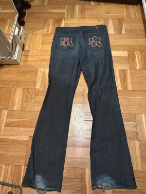 Rock & Republic mörkblå bootcut jeans - Mörkblå jeans från Rock & Republic med bootcut passform och snygga broderade bakfickor med glittriga detaljer. Klassisk femficksmodell med kopparfärgade nitar och knappar. Jeansen är gjorda i jeansmaterial och har en cool, lite utsvängd siluett.