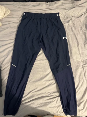 Mörkblå träningsbyxor Under Armour - Säljer ett par mörkblå träningsbyxor från Under Armour med elastisk midja och logga både fram och bak. Byxorna har smal passform, ventilerande paneler på baksidan och reflekterande detaljer vid benen. Perfekta för gymmet eller löpning.
