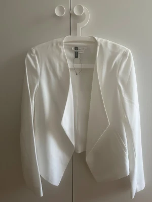 Vit kavaj med öppen front från H&M - Stilren vit kavaj från H&M med öppen front och snygg skuren nederkant. Långärmad modell i mjukt tyg som ger en clean och fräsch look. Perfekt för dig som vill ha en enkel men trendig kavaj till din outfit. Tyvärr har kavajen en liten defekt på armen