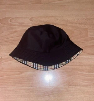  Svart bucket hat  - Snygg bucket hat med klassiskt Burberry-inspirerat rutmönster i beige, svart, vit och röd. Hatten är vändbar med ena sidan helsvart och andra sidan rutigt. Perfekt för att lyfta din streetwear-look och ge lite lyxig vibe. Materialet känns som bomull eller polyester.
