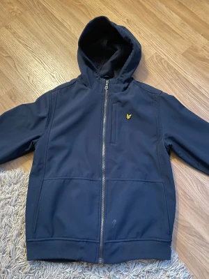 Mörkblå vindjacka från Lyle & Scott - Snygg mörkblå vindjacka från Lyle & Scott med huva och gul logga på bröstet. Jackan har dragkedja framtill, praktiska fickor och är fodrad med mjuk fleece på insidan. Perfekt för dig som vill ha en stilren och funktionell jacka.