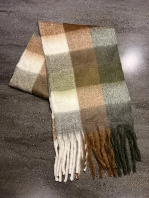 Halsduk - Mjuk och fluffig halsduk (som liknar acne studios) med rutigt mönster i vitt, grått, brunt och grönt. Långa fransar i ändarna ger en chill vibe. Perfekt att slänga över axlarna när det är kallt ute. Materialet känns som ull eller syntet och är riktigt skönt mot huden.