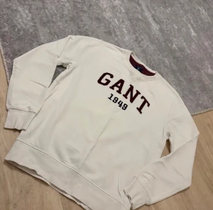 Vit sweatshirt från GANT med tryck - Vit sweatshirt från GANT med mörkrött tryck 'GANT 1949' på bröstet. Tröjan har rund hals med kontrastfärgad insida, långa ärmar och ribbade muddar vid ärmslut och nederkant. Perfekt för en chill och stilren look.