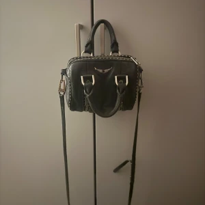 Svart Zadig & Voltaire handväska i skinn - Cool svart handväska från Zadig & Voltaire i mjukt skinn med silverfärgade detaljer och nitar längs kanterna. Väskan har både handtag och axelrem med logotyp, samt dragkedjor framtill och ett snyggt metallvingemblem. Perfekt storlek för det viktigaste.