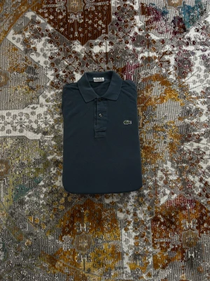 Blå långärmad pikétröja Lacoste - Blål ångärmad pikétröja från Lacoste med klassisk krage och knappar framtill. Tröjan har det ikoniska gröna krokodilemblem broderat på bröstet. Tillverkad i bomull med en stilren och tidlös look som funkar till många olika outfits.
