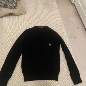 Svart v-ringad tröja från Lyle & Scott - Svart v-ringad tröja från Lyle & Scott med klassisk gul broderad logga på bröstet. Tröjan har lång ärm och är tillverkad i mjukt material som känns skönt mot huden. Perfekt för dig som gillar stilrena och enkla plagg med en snygg detalj.
