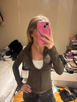 Mörkgrön slim träningsjacka med dragkedja - Säljer en mörkgrön träningsjacka med slim passform och hel dragkedja framtill. Jackan har en snygg krage och är långärmad, perfekt för dig som gillar en sportig och clean look. Materialet känns lätt och smidigt, vilket gör den enkel att bära till jeans eller leggings.