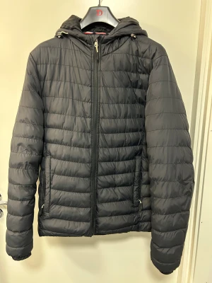 Svart pufferjacka från Vinson Polo Club - Svart pufferjacka från Vinson Polo Club med huva och dragkedja framtill. Jackan har quiltad design med horisontella sömmar och två fickor med dragkedja. Perfekt för kyliga dagar och har en clean, sportig look.