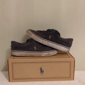 Mörkblå sneakers från Polo Ralph Lauren - Snygga mörkblå sneakers från Polo Ralph Lauren med klassisk låg profil och rund tå. Skorna har vit sula, snörning och Polo-logga i guld på sidan. Materialet är canvas och detaljerna är stilrena med en platt sula som ger en chill vibe ni får gärna fråga frågor