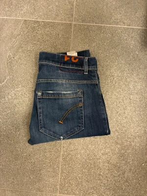 Blå skinny jeans från Dondup i modellen George  - Snygga blå jeans från Dondup i modellen George med skinny passform och klassisk femficksdesign. Jeansen har ljusa slitningar framtill och bak, samt kontrastsömmar i orange. Tillverkade i mjukt denim-material och har en läderpatch med Dondup-logga bak i midjan.