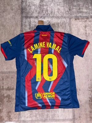 FC Barcelona Lamine Yamal #10 tröja - Säljer en FC Barcelona matchtröja med Lamine Yamal och nummer 10 på ryggen. Tröjan är kortärmad, har ett randigt mönster i blått och rött med gula detaljer och klubbmärke på bröstet. Tillverkad i polyester, med UNHCR och Spotify-logga tryckt på framsidan.
