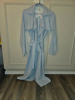 Ljusblå trenchcoat med bälte - Säljer en lång, ljusblå trenchcoat från Miss Selfridge. Jackan har en snygg oversized krage, långa ärmar och ett matchande bälte i midjan för extra stilpoäng. Perfekt för dig som vill ha en clean och trendig look under vår och höst.
