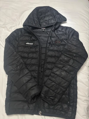 Svart dunjacka från Ellesse - Säljer en svart dunjacka från Ellesse i storlek S. Jackan har huva, quiltad design och dragkedja framtill. Ellesse-logga på bröstet och snygga detaljer. Perfekt för dig som vill ha en stilren och sportig look.