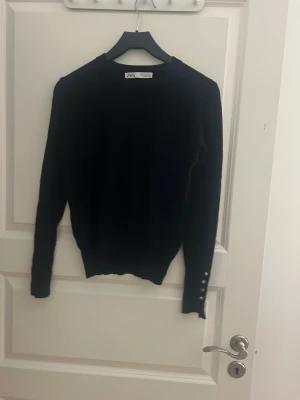 Svart långärmad tröja från Zara - Svart långärmad tröja från Zara med rund hals och snygga guldfärgade knappar på ärmsluten. Tröjan har en slim passform och är tillverkad i mjukt material som känns skönt mot huden. Perfekt för dig som gillar stilrena detaljer.
