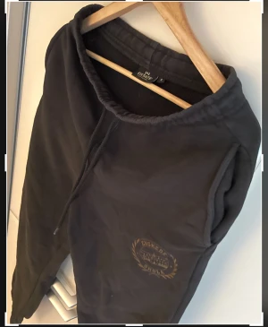 Svarta mjukisbyxor Estate S - Svarta mjukisbyxor från Estate Clothing med dragsko i midjan och tryck med texten 'Enskede Skola' på ena benet. Byxorna har resår i midjan och vid bensluten för en skön passform. Perfekta för chill eller träning.