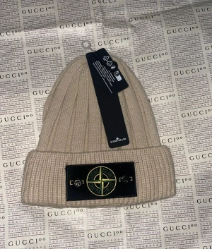 Beige ribbad mössa från Stone Island - Snygg beige ribbstickad mössa från Stone Island med klassisk logopatch framtill. Perfekt för dig som vill ha en clean och stilren look med streetkänsla. Mössan har uppvikt kant och är tillverkad i mjukt material som håller dig varm.