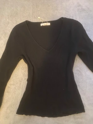 Svart ribbad tröja från Amisu - Svart långärmad tröja från Amisu med snygg v-ringning och ribbad struktur. Modellen är slim och sitter tajt mot kroppen, perfekt för dig som gillar en enkel men stilren look. Materialet är mjukt och stretchigt, vilket gör den skön att bära.