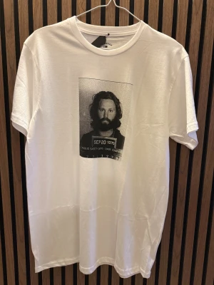 Vit t-shirt med tryck på Jim Morrison - Vit t-shirt från Fourstar i 100% bomull med ett svartvitt tryck av ett porträtt på bröstet. Klassisk rund halsringning och korta ärmar. Perfekt för dig som gillar streetwear och vill ha en cool statement-tee.