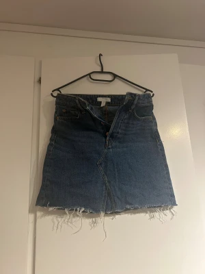 Mörkblå jeanskjol med rå kant - Säljer en mörkblå jeanskjol med klassisk femficksdesign och rå, fransig kant nedtill. Kjolen har en kort passform och är tillverkad i bomull. Perfekt för dig som gillar en avslappnad och cool stil.