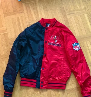 Röd och svart bomberjacka NFL Buccaneers - Säljer en fet bomberjacka från Pull&Bear x NFL med Tampa Bay Buccaneers tryck. Jackan är röd och svart, har ribbade muddar med svarta ränder, lång ärm och knappar framtill. Materialet är polyester och jackan har NFL patch på ärmen. Perfekt för dig som gillar amerikansk fotboll.