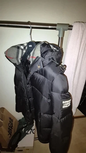 Svart Burberry pufferjacka med huva - Svart pufferjacka från Burberry med ikonisk rutig huva i grått, vitt och rött. Jackan har dragkedja och knappar framtill, två sidofickor med dragkedja och en patch med Burberry London England på ärmen. Perfekt för kalla dagar med sin tjocka vaddering. har inköpskvitto till den har inte använt så länge 