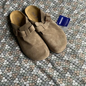 Birkenstocks - Säljer ett par snygga Birkenstock sandaler i beige mocka med rund tå och justerbart spänne i metall. Skorna har en platt sula och är perfekta för dig som gillar klassisk och avslappnad stil. Passar både till jeans och shorts.