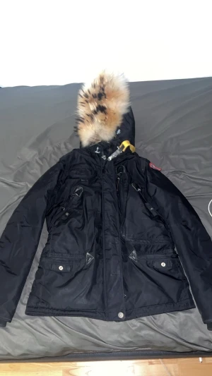 Svart Parajumpers vinterjacka med päls - Svart Parajumpers vinterjacka från Masterpiece Series, slim fit. Jackan har huva med fluffig pälsdetalj, flera fickor med knappar och dragkedja, samt den klassiska gula remmen vid kragen. Perfekt för kalla dagar och har snygga quiltade sömmar.