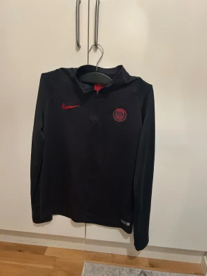 Svart PSG half zip tröja från Nike - Svart långärmad PSG tröja från Nike med röd logga och klubbmärke på bröstet. Tröjan har en half zip dragkedja och är gjord i mjukt polyester-material, perfekt för fotbollsträning. Snygg röd detalj på ryggen och sportig passform.