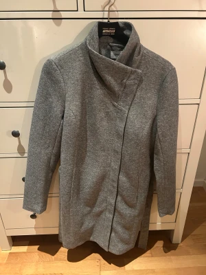 Grå kappa från ONLY med hög krage - Säljer en stilren grå kappa från ONLY med hög krage och asymmetrisk dragkedja. Jackan har lång ärm och en rak, clean siluett som ger en modern vibe. Perfekt för dig som gillar minimalistisk design och vill ha något som funkar hela hösten och vintern.