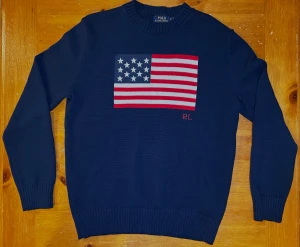 Ralph Lauren USA Flagga - Snygg marinblå stickad tröja från Polo Ralph Lauren med amerikansk flagga broderad på bröstet och RL-detalj. Tröjan har rund halsringning, ribbade muddar och är långärmad. Perfekt för dig som gillar klassisk och preppy stil. Skick: 9/10. Storlek M men sitter mer som L!