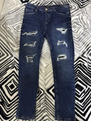 Blå ripped jeans från Amiri - Fejk amiri Säljer ett par mörkblå jeans från Amiri med flera slitningar och hål på framsidan dom e stängda med tyg. Klassisk femficksmodell med knappgylf och snygga detaljer. Jeansen har en slim passform och är tillverkade i jeansmaterial med cool tvättad look.