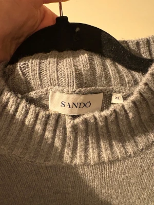 Grå stickad tröja från Sandö - Säljer en grå stickad tröja från Sandö i storlek XL. Tröjan har en ribbad hög hals och ribbade muddar vid ärmslut och nederkant. Perfekt för dig som gillar enkel och clean stil med lite loose passform. Aldrig använd.