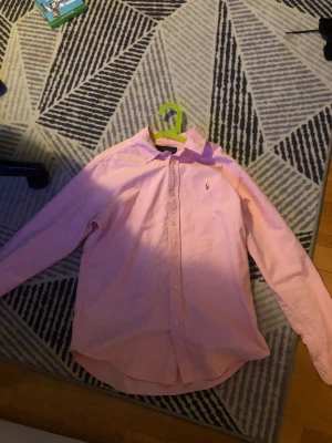 Rosa skjorta från Ralph Lauren - Snygg rosa skjorta från Ralph Lauren med klassisk krage och knäppning framtill. Skjortan har lång ärm och ikonisk broderad logga på bröstet. Tillverkad i mjuk bomull som känns skön mot huden. Perfekt för dig som gillar stilrena och tidlösa plagg.