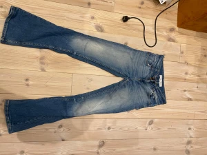 Blå bootcut jeans från Bershka - Snygga blå jeans från Bershka med bootcut passform och klassisk femficksdesign. Jeansen har ljusa slitningar framtill och är tillverkade i jeansmaterial med orangea sömmar. Perfekt för dig som gillar retrovibbar och vill ha ett par jeans med lite extra attityd.