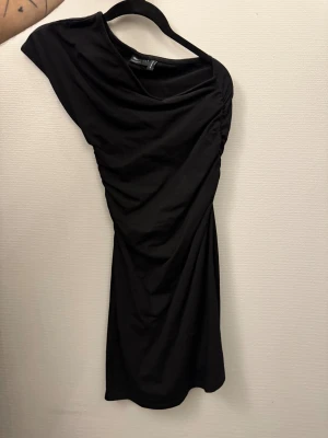 Svart oneshoulder kortklänning - Säljer en svart oneshoulder klänning från ASOS. Klänningen har draperad effekt och är figurnära med kort längd. Perfekt för dig som gillar en stilren och trendig look. Materialet är mjukt och stretchigt, vilket gör den bekväm att bära.