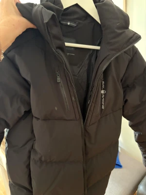 Svart Sail Racing pufferjacka med huva - Svart pufferjacka från Sail Racing med stor huva och två bröstfickor med dragkedja. Jackan har en stilren look med logga på bröstet och huvan. Perfekt för kalla dagar och har en schysst, lite loose passform.