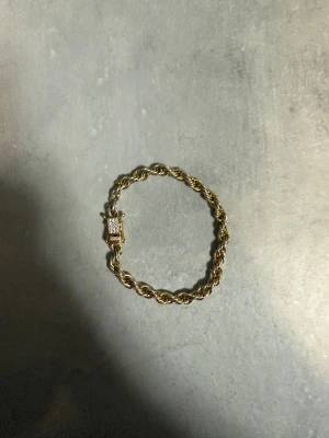 Guldarmband med glittrig låsdetalj - Snyggt guldarmband med tvinnad kedja och en låsdetalj som är dekorerad med små glittrande stenar. Armbandet har en klassisk och stilren look som passar perfekt till din outfit. Ingen synlig märkning på armbandet.