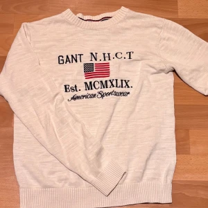 Vit stickad tröja från GANT - Vit stickad tröja från GANT med broderad amerikansk flagga och text på bröstet. Rund hals med ribbad kant och långa ärmar. Snygg sportig stil med klassiska detaljer och mjukt material som passar perfekt till jeans.