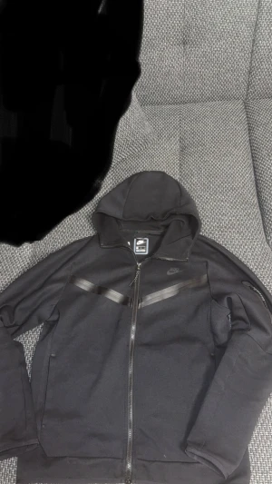 Svart vindjacka från Nike med huva - Svart Nike vindjacka med huva och hel dragkedja framtill. Snygga svarta detaljer i form av diagonala ränder över bröstet och en ficka med dragkedja på ärmen. Jackan har en sportig look och är perfekt för dig som gillar streetwear. PRIS KAN DISKUTERAS!