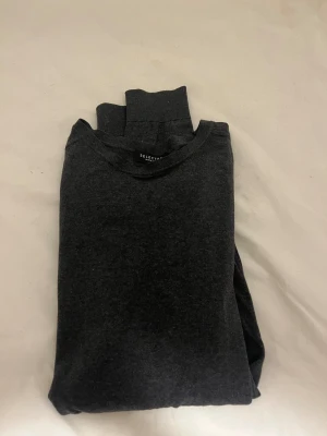 Mörkgrå långärmad tröja SELECTED - En stilren mörkgrå långärmad tröja från SELECTED med rund hals och ribbade muddar vid ärmslut och nederkant. Tröjan är tillverkad i mjuk bomull och har en klassisk, normal passform som funkar till många olika looks.